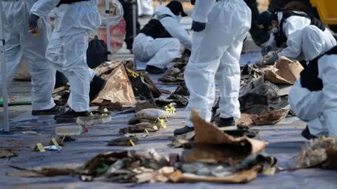Getty Images A row of debris from Jeju Air Flight 2216 laid on the ground, surrounded by investigators all wearing white, shot from their waists down