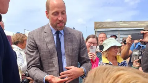 Prince William, dressed in a grey patterned suit with a light blue shirt and navy blue tie with his hand out accepting a crisp. There are people in the background taking photos on their phones.
