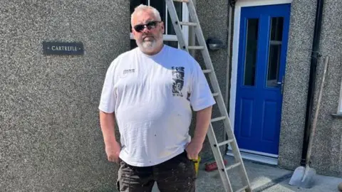 Jason is wearing a white t-shirt and black shorts. He has grey hair and beard and is wearing black sunglasses. He is stood in front of a pebble-dashed house. It has a blue door and a ladder and shovel in view behind him.