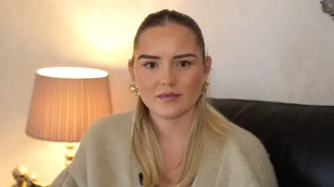 John Fairhall/BBC A young woman with long blonde hair smiles at the camera. She has dark eyebrows and a nose ring. She is wearing a light-coloured top and is sitting in front of a pale background.