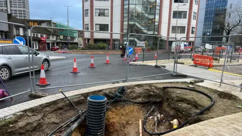 A view of a road in Swindon where you can see a hole in the road, pipes and cables poking out and fencing behind it. There are cones are cars driving around.