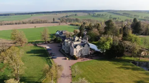 Graham and Sibbald Shieldhill Castle is a three storey white modern manor sitting in a lush green setting with a long red driveway