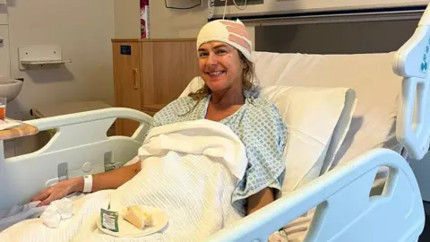 Joanna James A woman in a hospital bed with her head bandaged, she is wearing a hospital gown and smiling. 