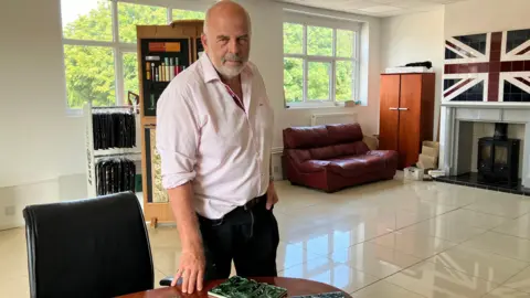 A man in a pink shirt and dark jeans has one hand leaning on a table. Behind him is a tiled floor, windows looking onto trees, a leather settee and a fireplace with a tiled union jack 