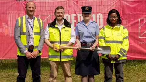 Two men and a woman wearing yellow hi-vis jackets stand either side of a woman wearing an RAF uniform - a dark blue hat, a light blue shirt and grey skirt. The woman in the RAF uniform is holding a shovel sideways. A big red banner has been draped over a metal fence behind them.