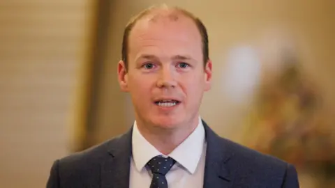 PA Media A man with balding dark hair, a white shirt dotty tie and navy suit speaks 