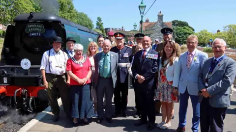 Andrew PM Wright A group of people including a woman in mayoral chains and two men in military uniforms with medals standing on a station platform with a steam locomotive in the background