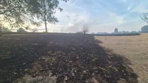 Staffordshire Fire and Rescue Service A large area of grass scorched black by fire in a park. Trees can be seen in the area, with buildings and goal posts on the horizon.