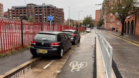 Meghan Hoyt Cars parked inside cycle lane with gate to the right.