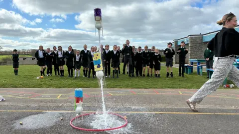 A teacher is running away after setting a rocket experiment in a school playing field. There are about 20 pupils in uniform watching, some are laughing and excited. The experiment is three plastic bottles filled with a white liquid which have exploded in to the air like rockets.