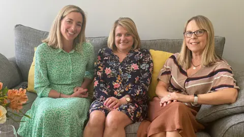 Three women sit side by side on a grey sofa. They all have straight blonde hair which is around their shoulders. They are smiling and each have their hands in their lap. There are two glasses of water and a bouquet of flowers on a coffee table in front of them.