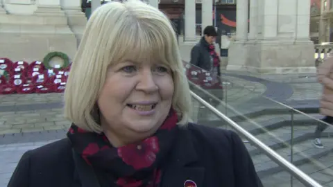 Carol has a blond bob. She's wearing a scarf with a floral design and a jacket. She's standing in the grounds of city hall with wreaths in the background.