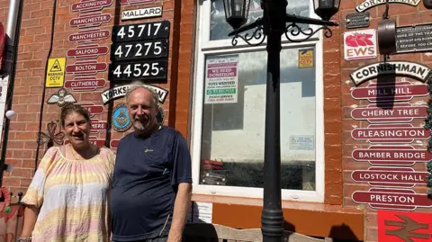 Eric and Wendy Coupe standing in the sunshine outside their red brick terraced house covered in vintage railway signs