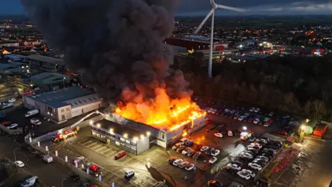 A fire is blazing from a building in an industrial estate. In there car park there are a large number of cars. One fire engine appears to be tackling the blaze 