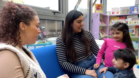 The actor sits on a sofa talking to one mum while two pre-school children watch her intensely.