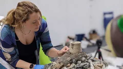 Jamie Willow Jamie leans over a sculpture she is working on which has stones and a tower on it. She is wearing a blue plaid shirt with a black top under it. She has strawberry blond hair tied into a ponytail.