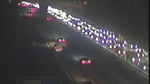 National Highways Cars on a carriageway. Their headlights are bright as it is dark and they are lined up three-deep in traffic. The opposite carriageway is clear with cars moving freely.