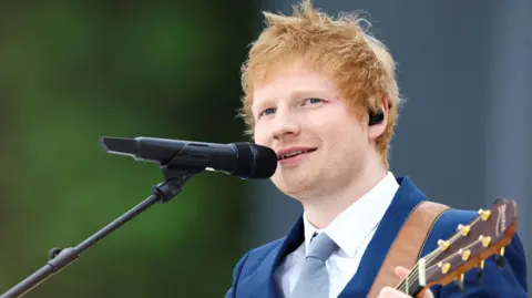 Ed Sheeran a man who is standing outside, wearing a navy suit with white shirt and grey tie. He is singing into a microphone and holding a wooden guitar. 