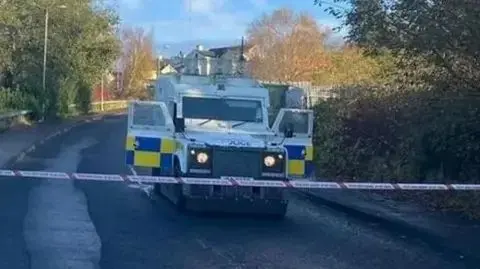 The picture shows an armoured police land rover with both doors open stopped on a road behind police tape after a bomb attack