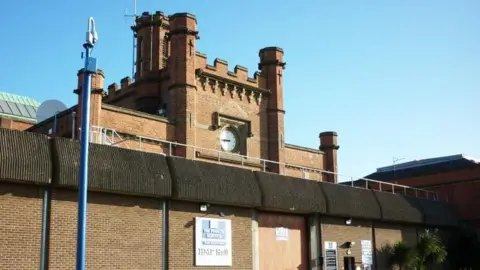 The main gate of Hull Prison it has a large wall topped with concrete barriers and a central tower with turrets and a clock