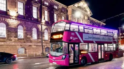A purple double-decker bus drives past a large cream building at night. 