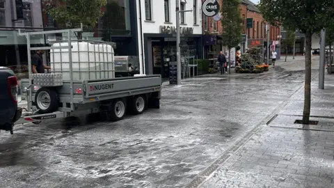 A black paved street with businesses down the side of it. There are suds on the ground as people 
