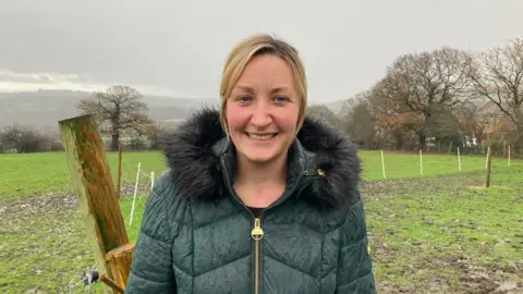 A woman with blond hair wearing a green hooded coat standing in a field with trees in the background 