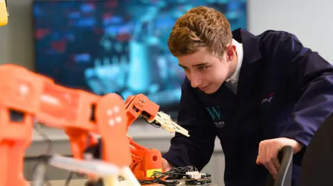 A young man is looking intently at a piece of orange machinery in front of him on a desk. He is wearing a navy uniform and leaning in over a chairback to look at the machinery.