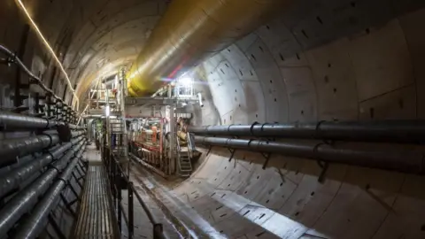 A view inside the Bromford Tunnel where lots of large pipes can be seen as well as crew in orange overalls