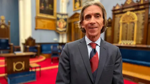 Tim smiles at the camera inside the Freemason Temple, He is wearing a grey suit with a light blue shirt and a red and dark blue tie. He has grey hair which is parted in the middle. Behind him - there are wooden boards on the walls with the names of Freemasons on  and flags with symbols and paintings of former leaders.