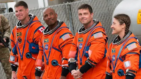 Getty Images Four astronauts wearing orange Nasa spacesuits, there are three men and one woman on the right. She has brown hair in two plaits. Both men on the far left and second from the right have short brown hair. The second man is bald. 