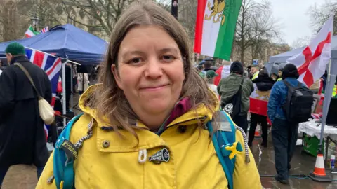Rachel Bee is wearing a yellow jacket and standing on a square with people behind her waving Iranian flags.