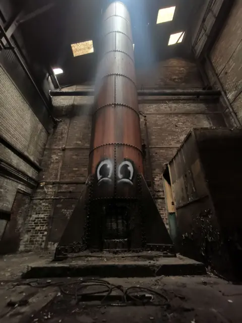 Psa Two large eyes painted on a large red or orange chute running into a disused building