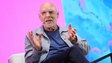 Getty Images Brian Eno sitting on a chair talking at a conference. He wears a grey blazer over a blue t-shirt. The background screen is bright pink and blue.