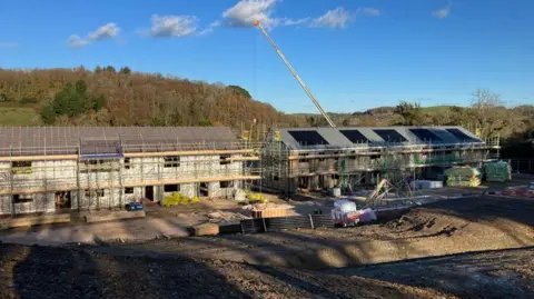 Blue skies and trees surround a building site where timber frame homes topped with solar panels can be seen covered with scaffolding.