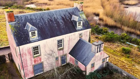 Jay Curtis Close up drone shot of one of the larger houses in the village. It is a white bricked property, with an extension on the behind. It is derelict with red marks on the sides and surrounds greenery and a lake.