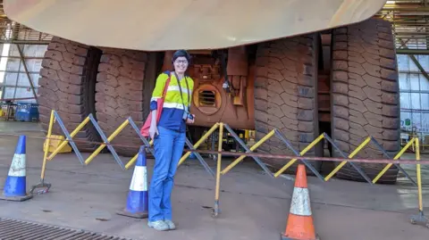 Zoe Corbyn Zoe standing next to a mining truck. The wheels look taller than him.