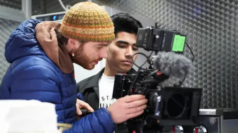 Two men are looking intently into the view finder of a camera. The man on the left, who has a beard, is wearing a blue jacket and a woolen hat. The other man is one of the students. He has dark hair, a black jacket and white T shirt.