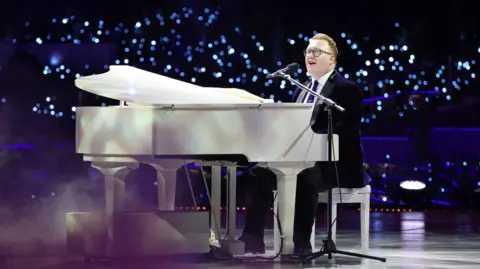 Getty Images A man in a suit singing on stage while sat behind a large white piano