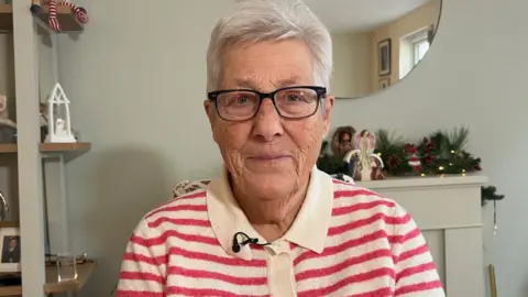 Mary Osmond is wearing a red and white striped top and looking at the camera. She is sitting in her living room. There is a mirror behind her and there are Christmas decorations in the room.