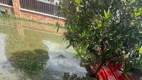 Supplied Waterlogged backyard garden with muddy standing water covering artificial grass and paving, a small tree growing at an angle, red plastic barriers stacked beside it, and a fence and house visible in the background.