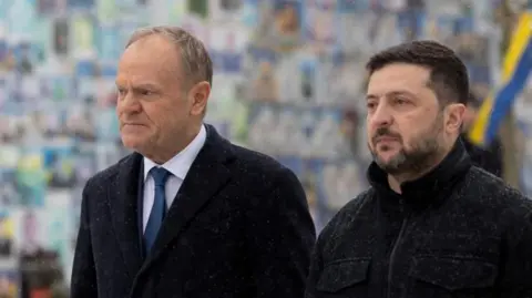 Polish Prime Minister Donald Tusk, an older man with short, blond hair and a dark overcoat, alongside Ukraine's Volodymyr Zelensky, a younger man with short, dark hair and beard, wearing a dark jacket. The pair are standing side by side.