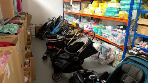 A room with shelves on the left and right. There are boxes and products such as nappies stacked on the shelves, and a line of black baby prams down the middle of the two sets of shelves
