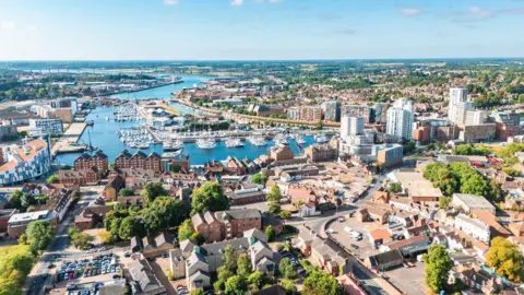 Chunyip Wong/Getty Images An aerial image of Ipswich showing the quay surrounded by residential buildings and trees. The sky is blue with few clouds in it.