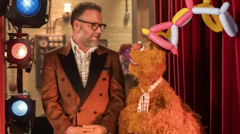 Disney Seth Rogen wears a 70s style smoking jacket and ruffled shirt as he stands with his hands folded in front of him next to studio lights - he is talking to puppet Fozzie, who wears an elaborate balloon hat 