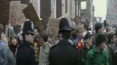 A crowd of protesting miners holding signs, with two policemen watching.