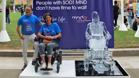 A man in a blue tshirt and white trousers standing outdoors next to a woman with dark hair and a blue tshirt in a wheelchair on a platform with a blue background and an ice sculpture of a person in a wheelchair