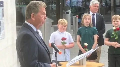 James Nicolson's nephew speaking at the unveiling of the plaque. Several children are standing beside him, listening. The project leader is behind them.