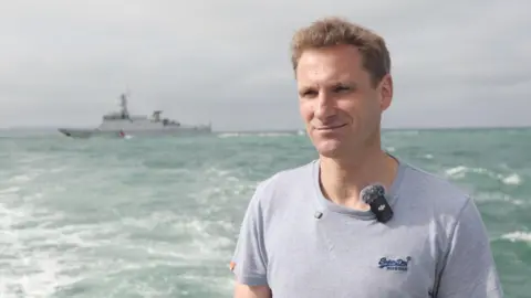 MP Chris Philip, in a grey t-shirt, stands on a boat in the sea. A large, grey ship is visible in the background.