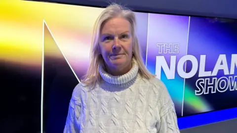 Sonya McMullan is looking into the camera. She's standing in a studio with a poster for the Nolan Show behind her on a screen.